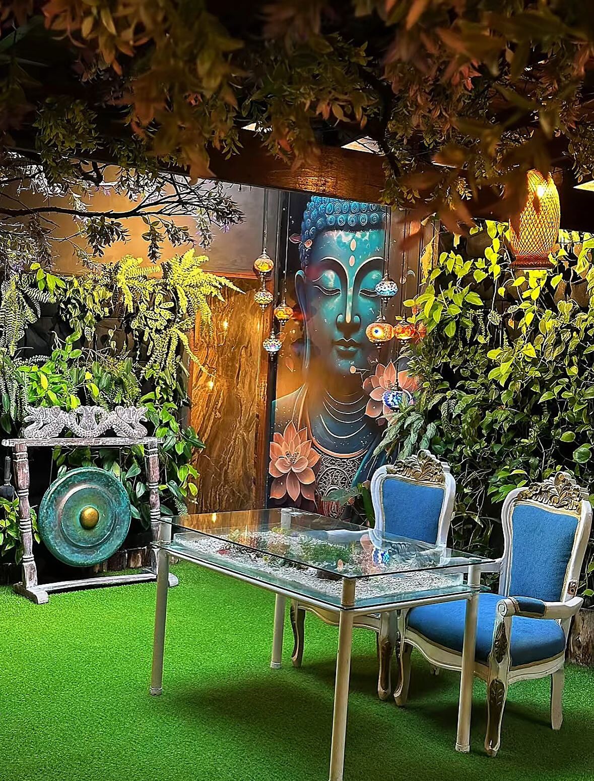Mesa de vidrio con dos sillas azules frente al mural del Buda, junto a un gong, entre plantas y ramas