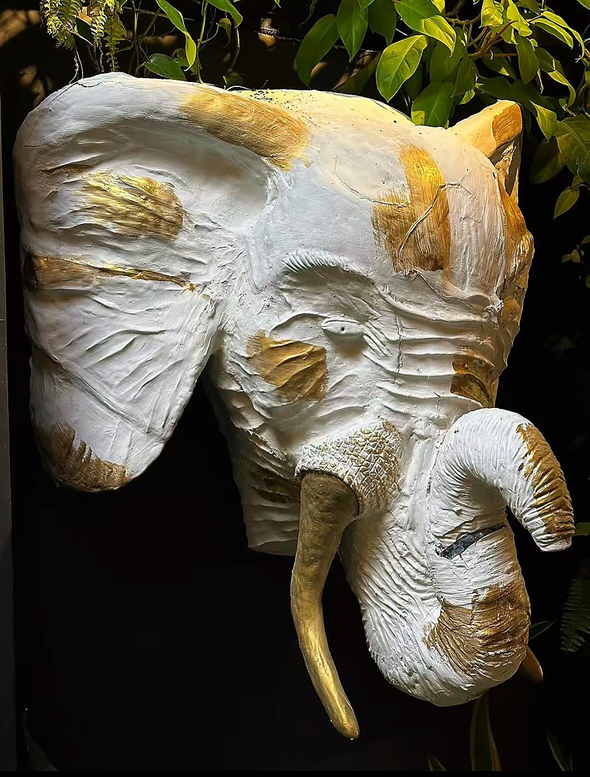 Cabeza de elefante blanca con detalles dorados colgada de la pared, entre hojas verdes