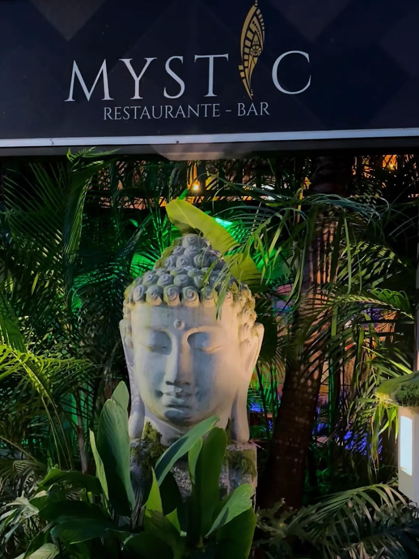 Cabeza de Buda en piedra entre palmas y plantas, bajo el letrero de Mystic Restaurante Bar, en la entrada
