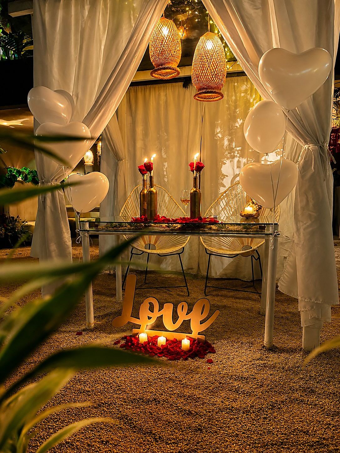 Mesa para dos en una cabaña de cortinas blancas sobre la arena, con velas, rosas, globos de corazón y un letrero Love