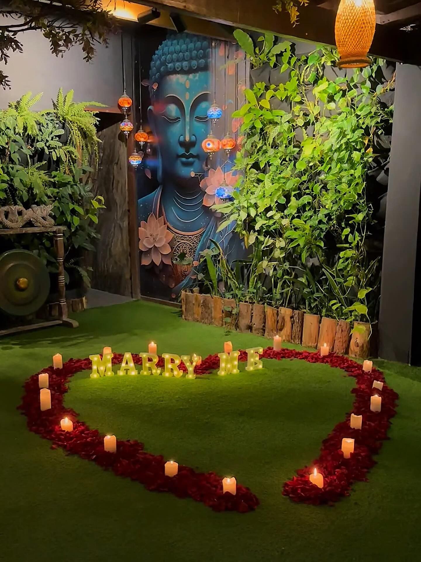 Pedida de mano en el Bosque Isabelino: corazón de pétalos y velas sobre el pasto, con el letrero Marry Me frente al mural del Buda azul