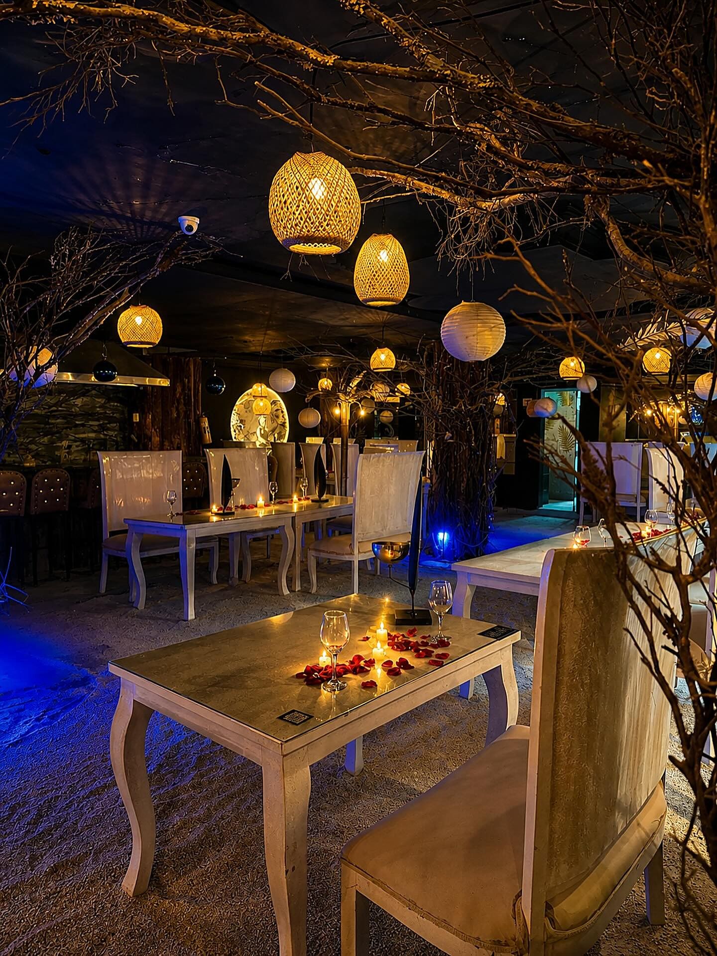 Salón de Playa Nocturna con mesas blancas, velas y pétalos sobre la arena, bajo ramas, faroles y una luz azul, con la luna al fondo