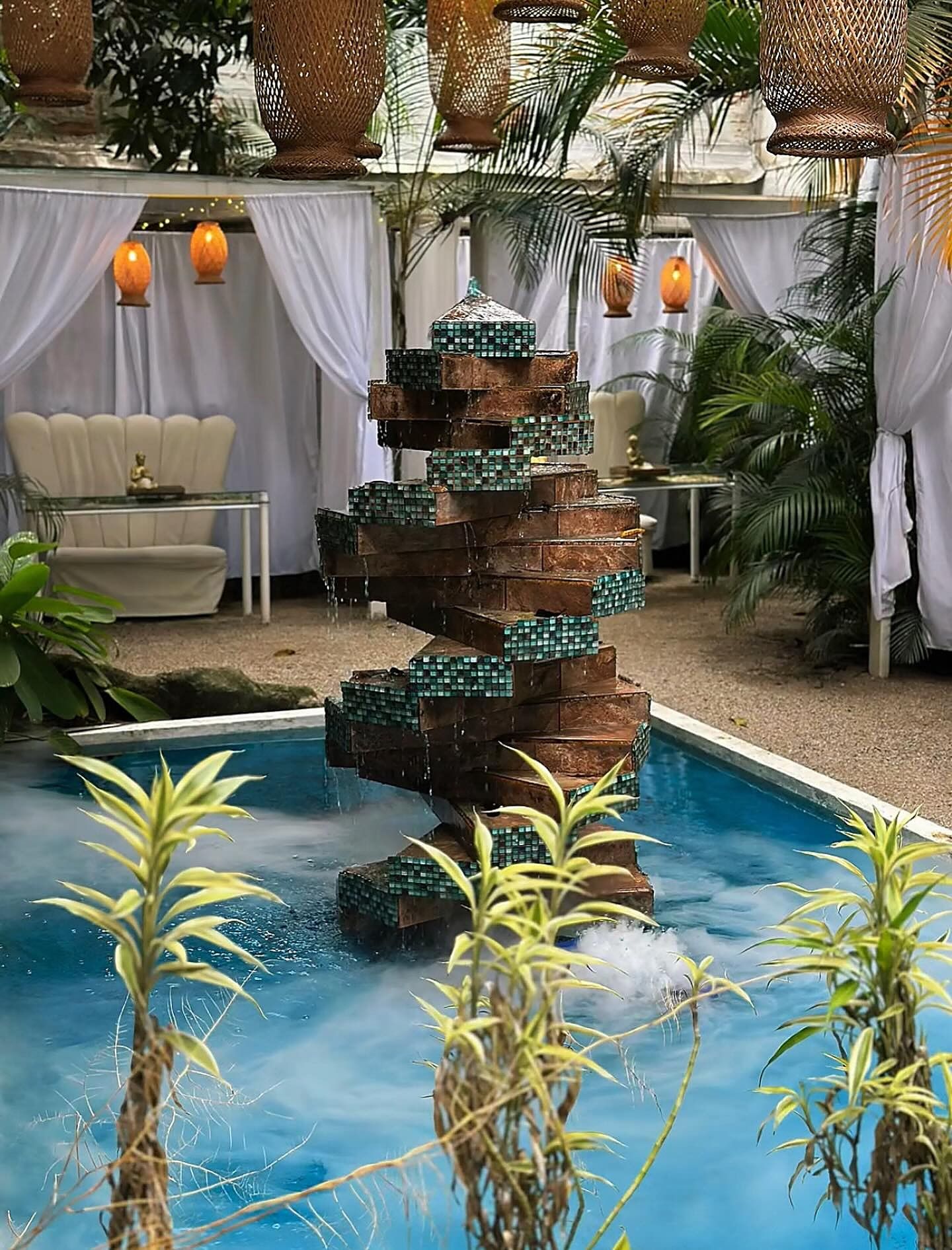 La fuente escalonada de Mystic sobre agua azul con neblina, con cabañas de cortinas blancas y linternas al fondo