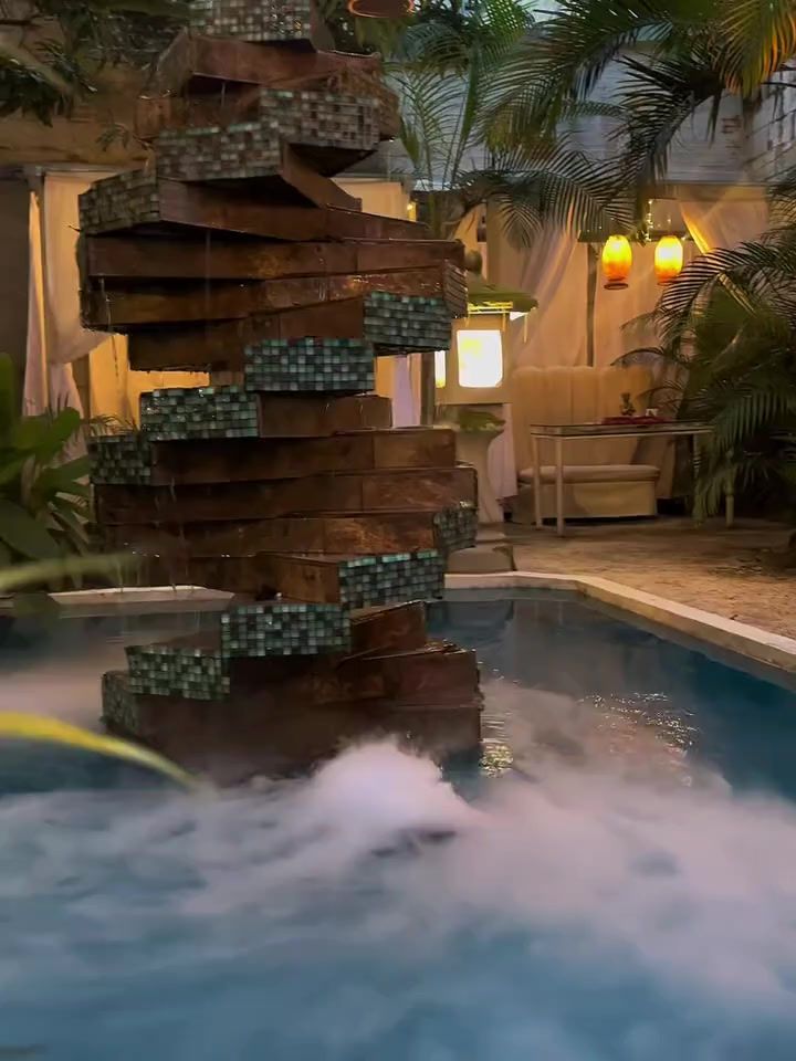 La fuente de Playa Oasis en Mystic, con neblina sobre el agua, una cabaña iluminada al fondo y linternas encendidas